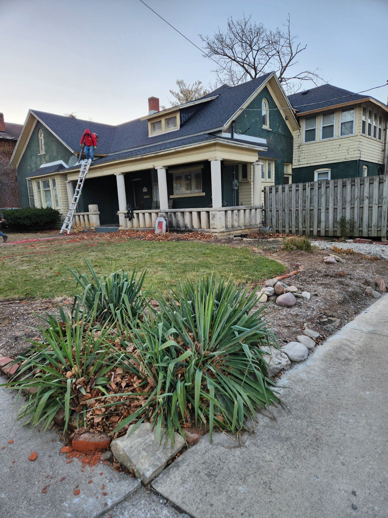 A person on a ladder repairs a green house with a porch and a yard, daytime.