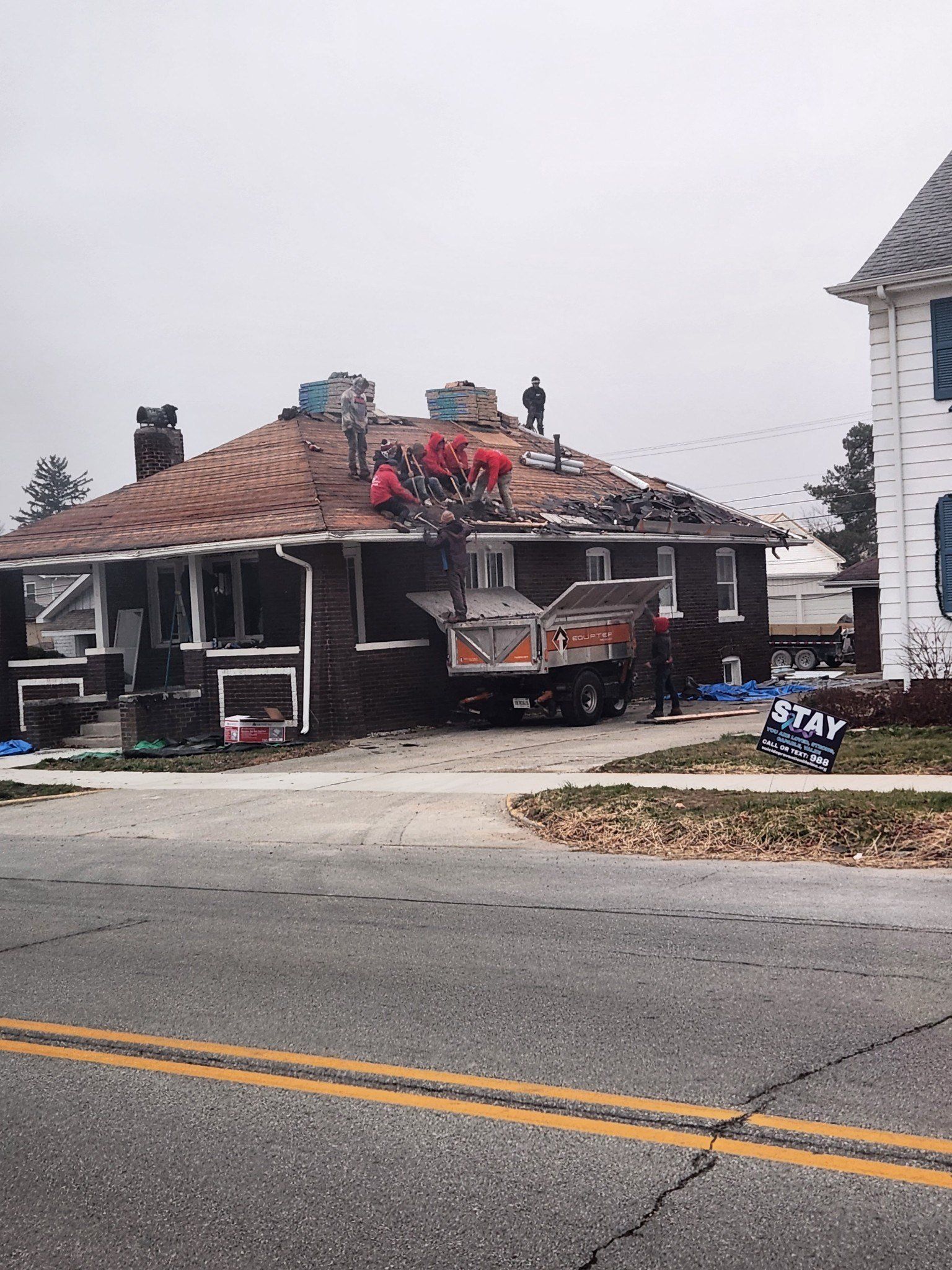 Roofers working on a brown roof. A truck and house are in the scene. Road and sign in front of the house.