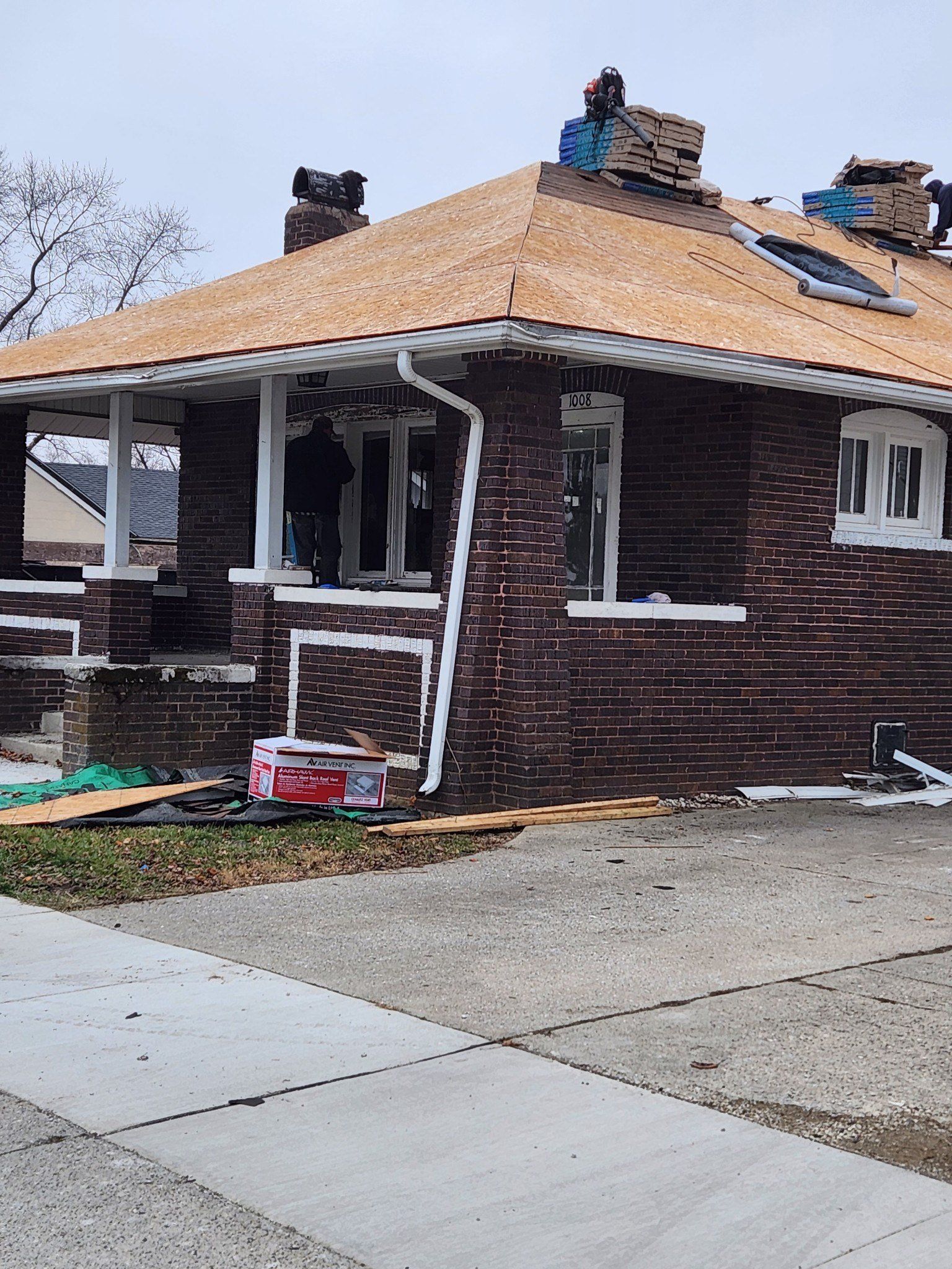 Brick house under construction; person in window; unfinished roof; gray sidewalk.