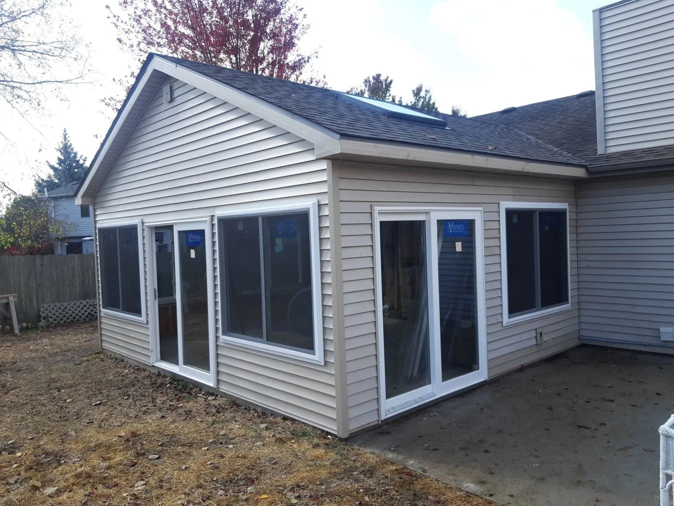 Sunroom addition on a beige house with large windows, a gray patio, and a glimpse of a fenced yard.