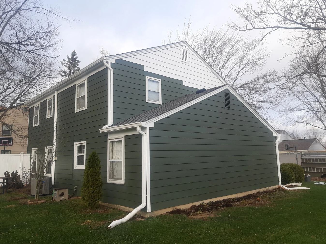 Two-story house with dark green siding, white trim, and a light roof, set in a yard with trees and a cloudy sky.