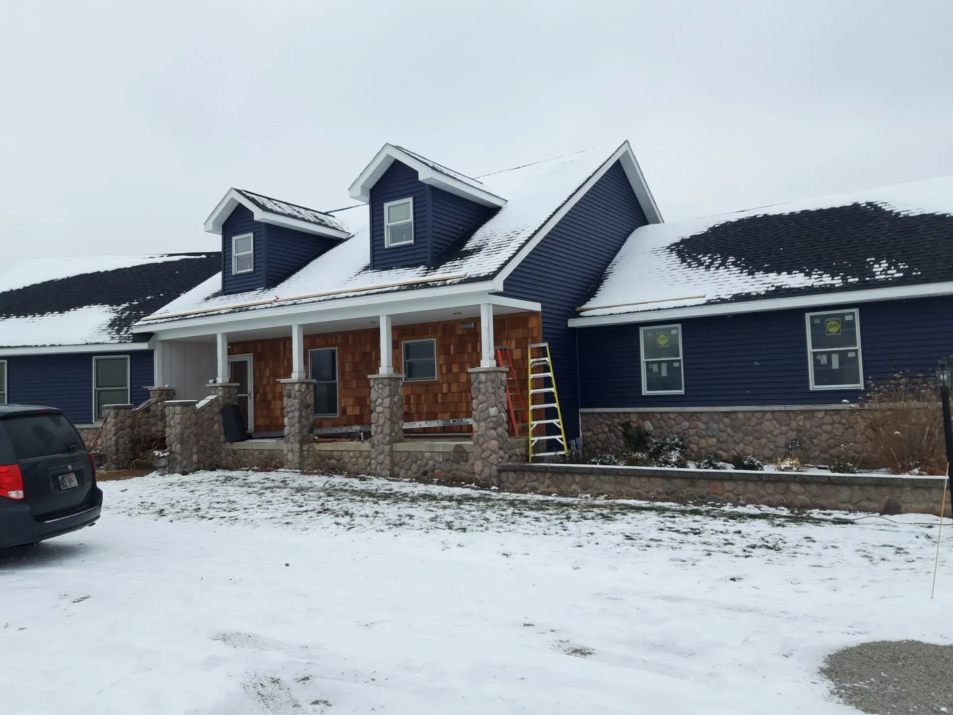 House with blue siding, brown brick columns, and snow on roof. A ladder leans against the house.