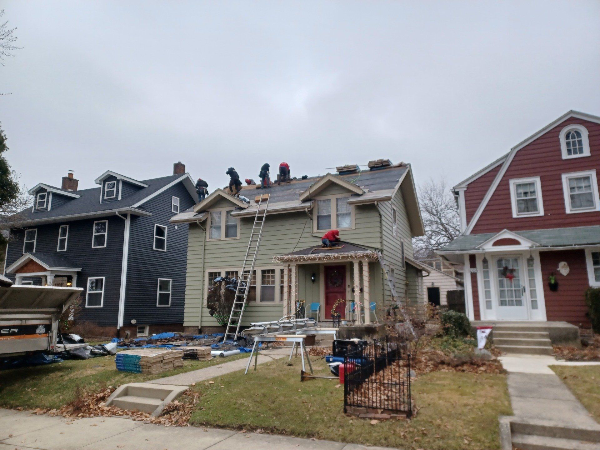 Workers on a roof replacing shingles on a light green house, with other houses visible.