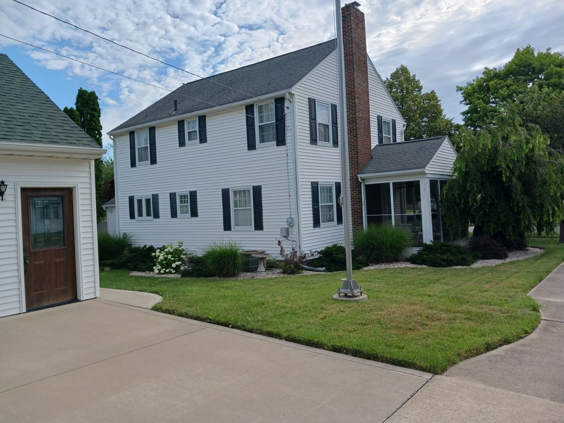 White two-story house with dark shutters, a chimney, and a screened-in porch, adjacent to a garage, on a grassy lawn.