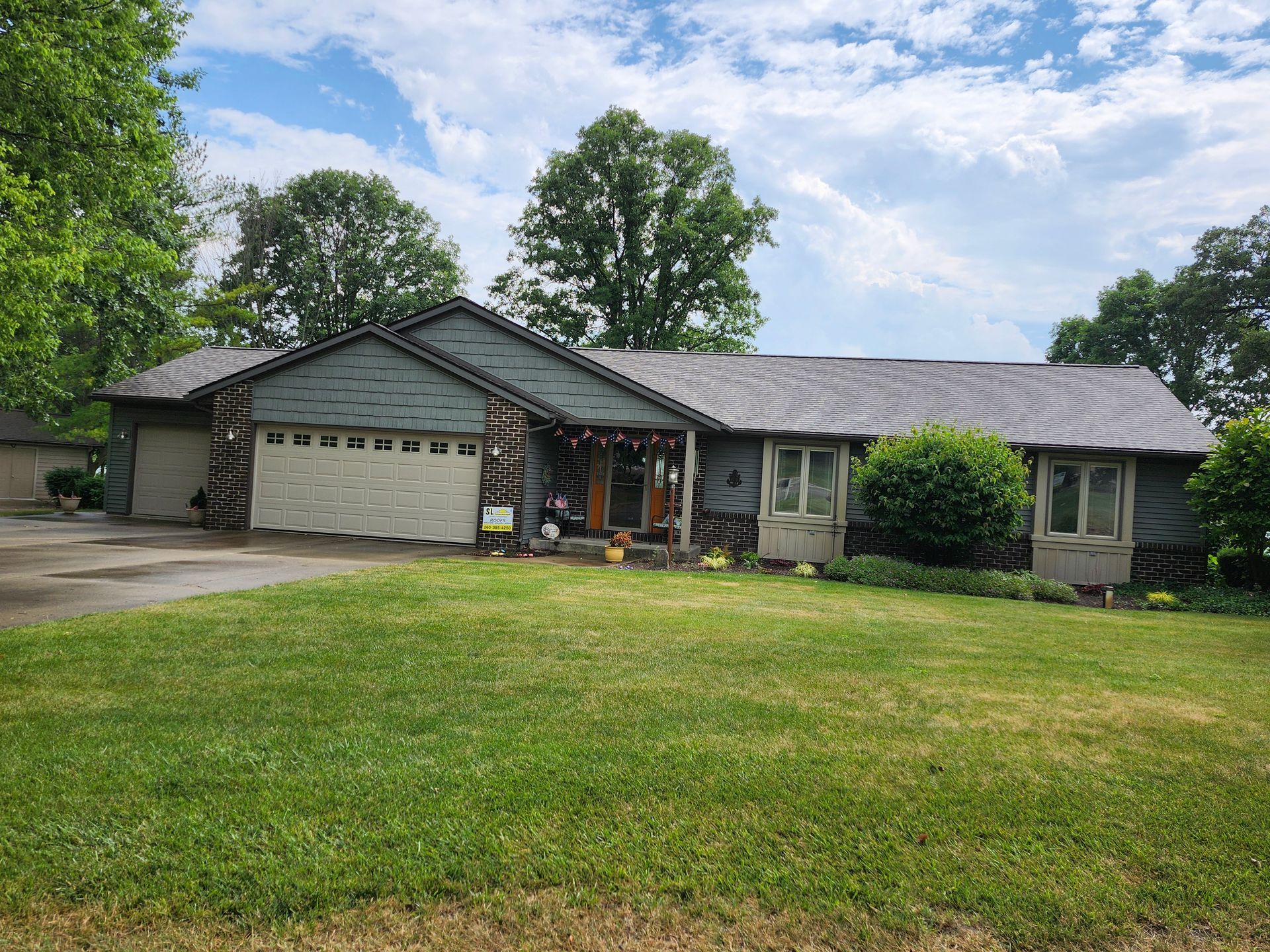 Ranch house with a gray roof, white garage doors, and a green lawn under a partly cloudy sky.