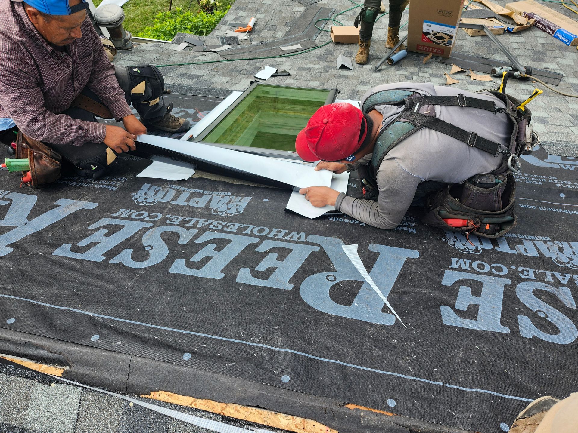 Two roofers installing flashing around a skylight on a roof covered with black underlayment.