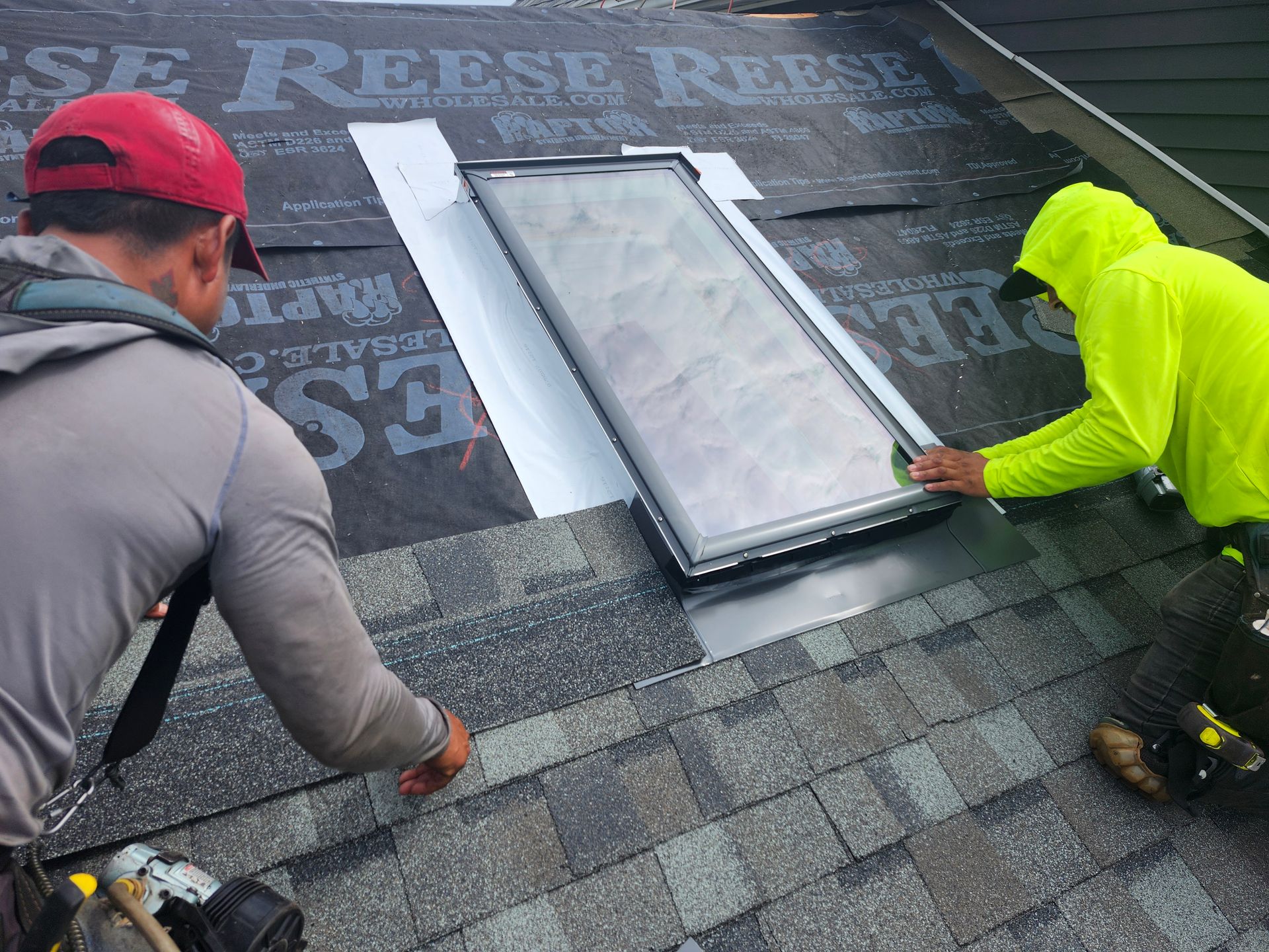 Two roofers installing a skylight on a shingled roof, using flashing materials.
