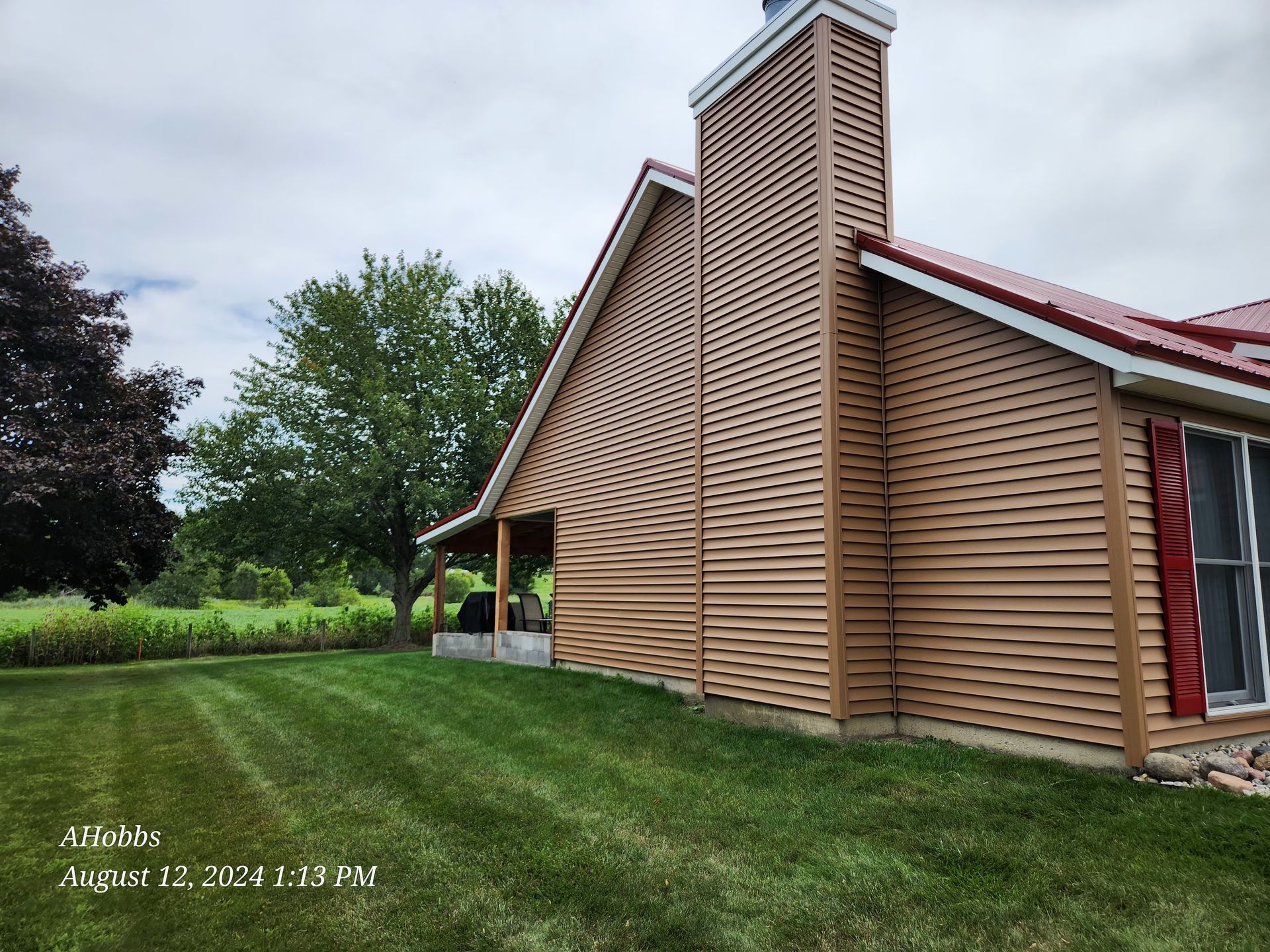House exterior with tan siding, red roof and trim, chimney. Green lawn and trees in background.