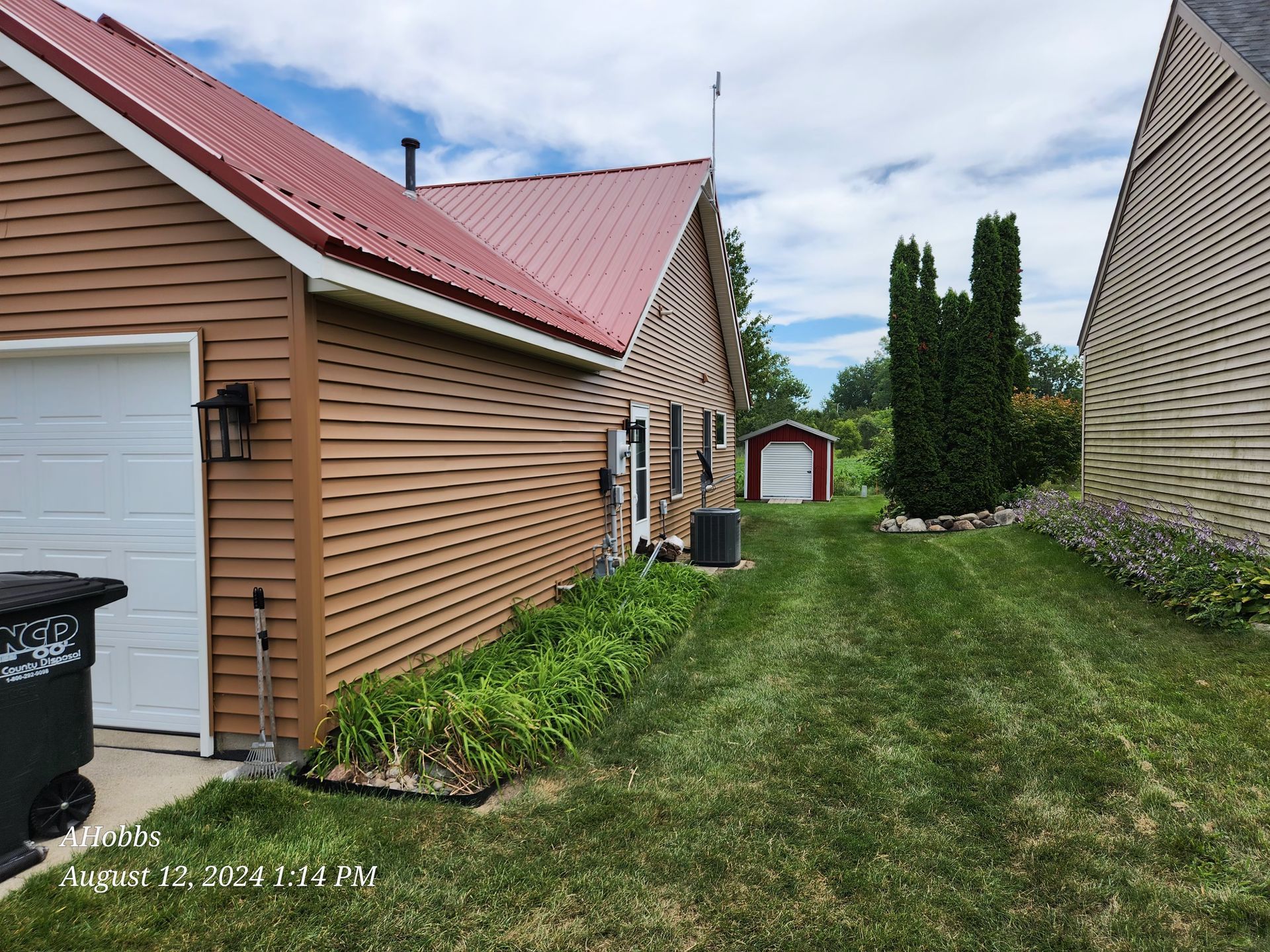Brown house with red roof, green lawn, small red shed in the distance, blue sky with clouds.
