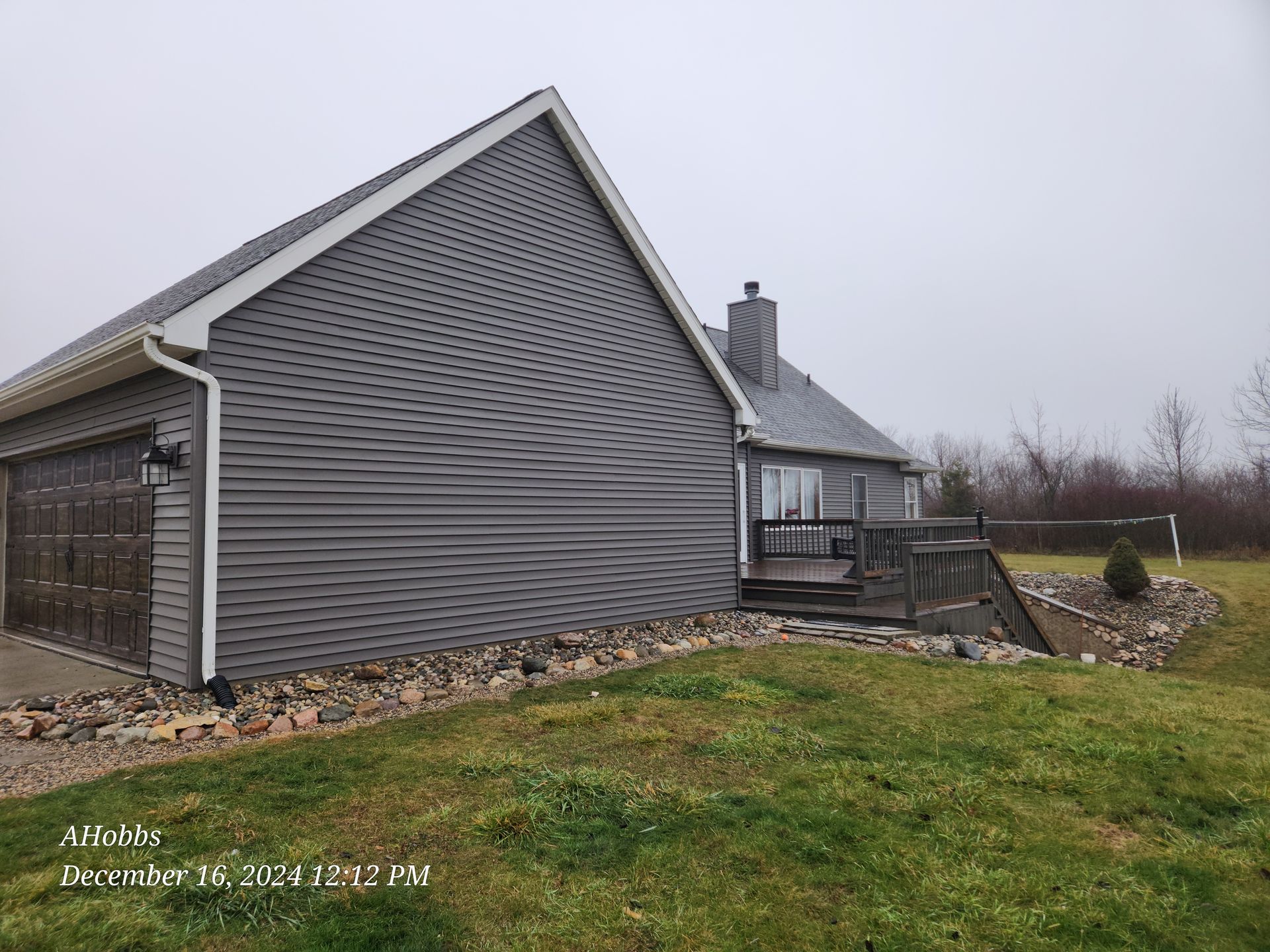 Gray-sided house with a garage, deck, and chimney, set on a grassy lot under a cloudy sky.