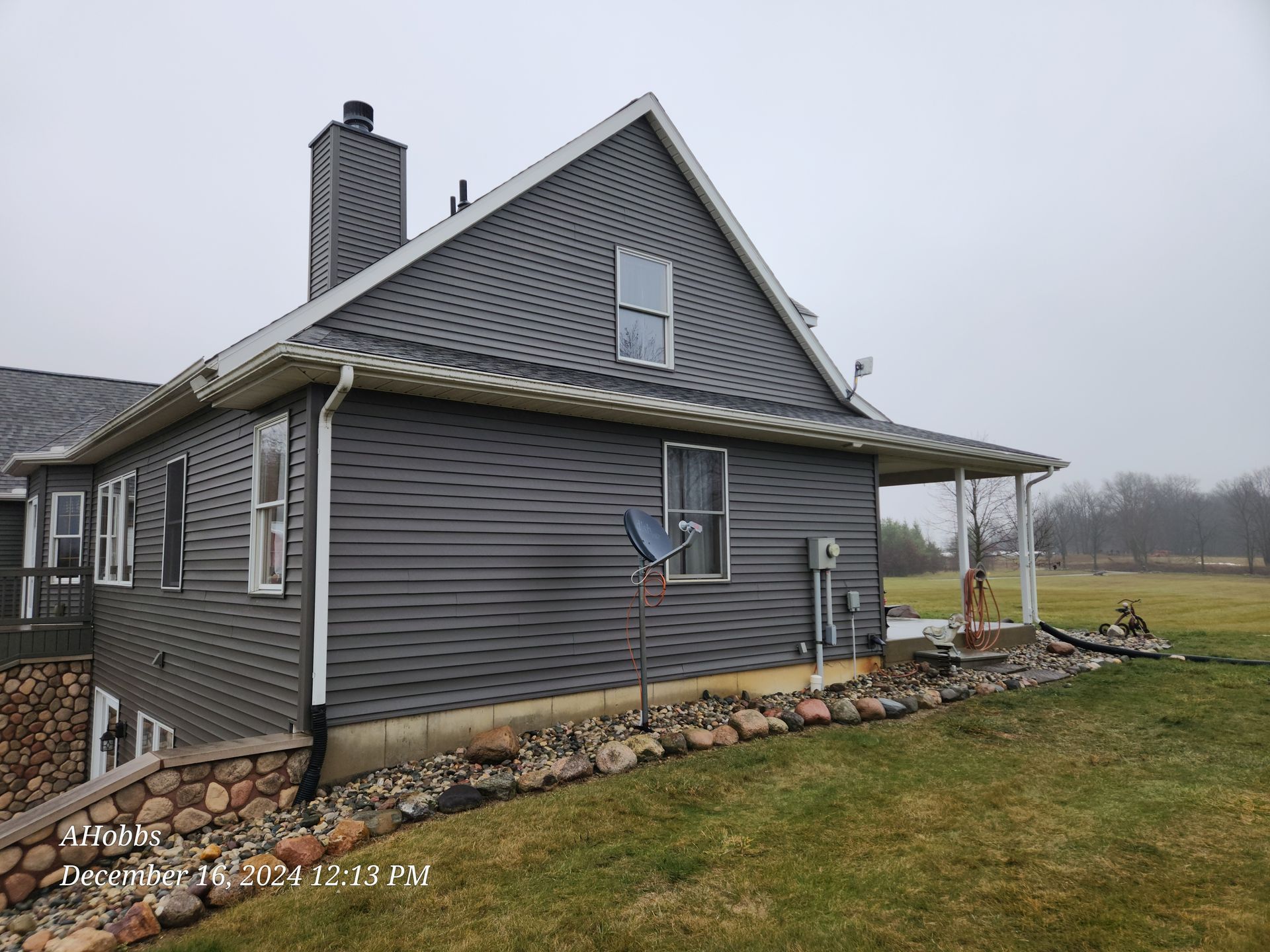 Gray house with stone retaining wall, lawn, and a cloudy sky.