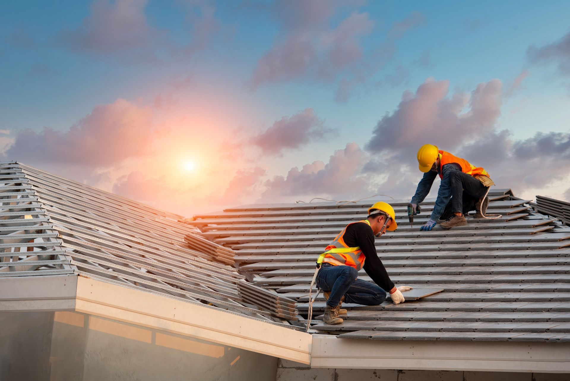 Two roofers working on a gray tiled roof under a partly cloudy sky at sunset.