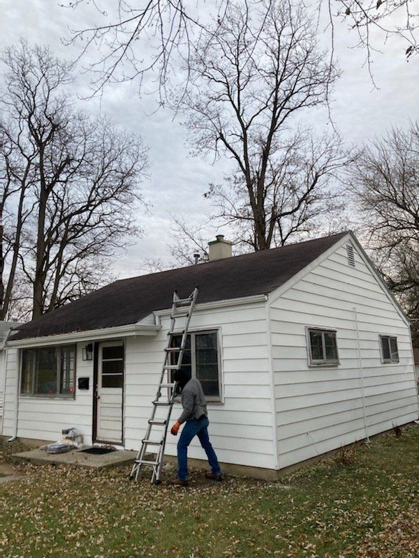 A person on a ladder by a white house with a dark roof, trees in the background.