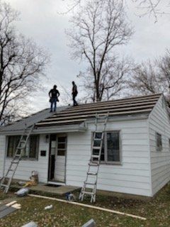Two roofers working on a white house roof, with ladders and bare trees in the background.