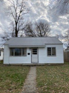 White one-story house with metal roof, small front porch and walkway. Overcast sky.
