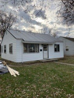 White house with metal roof and small porch, grass yard, cloudy sky.