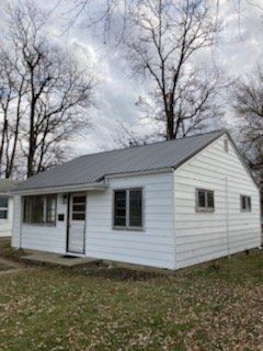 White, single-story house with metal roof, small windows, and bare trees in the background on a cloudy day.