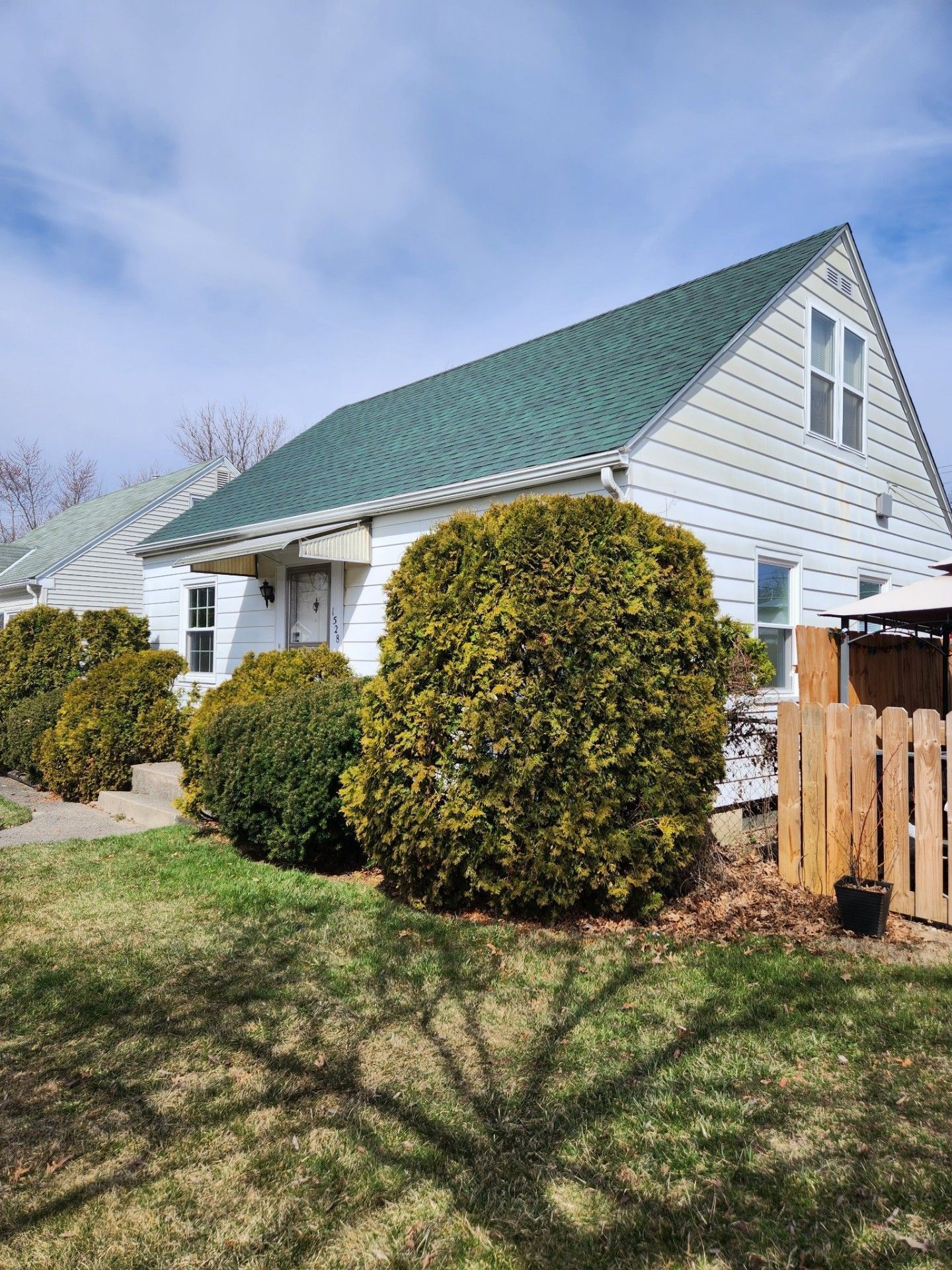 White house with green roof, large green bushes in front, wooden fence to the right.