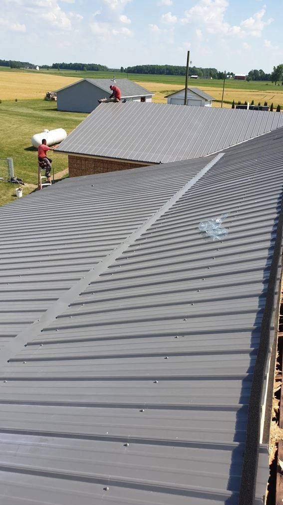 Two workers installing a gray metal roof on a rural building under a sunny sky.