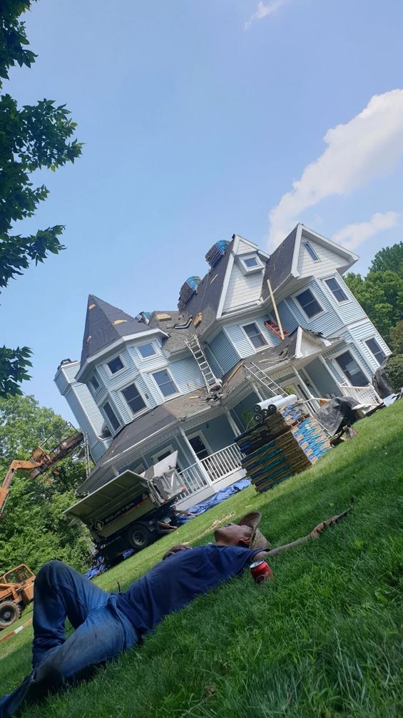 Man reclining on grass in front of a large, light-blue house under construction on a sunny day.