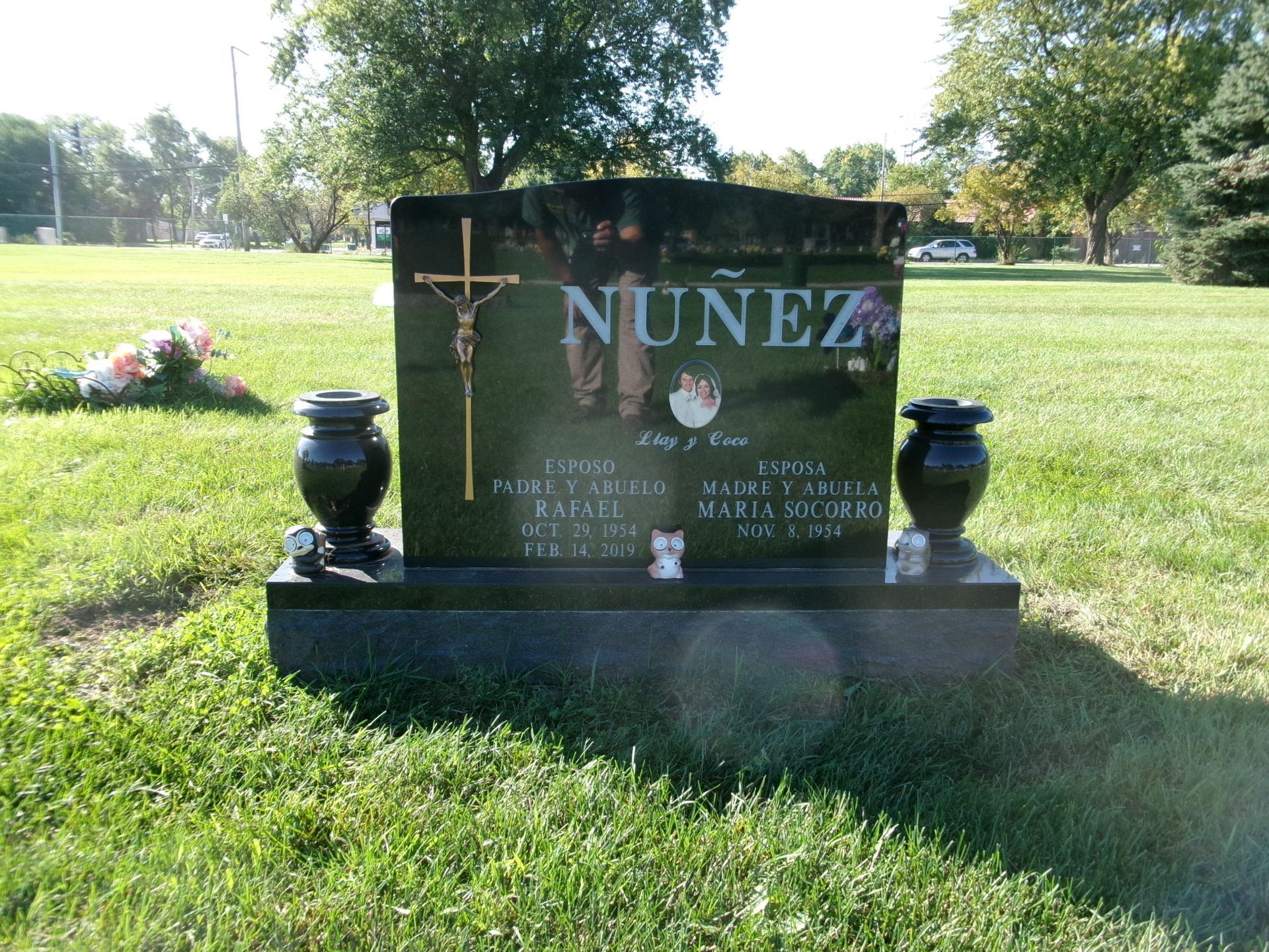 A black granite Nunez family headstone with a gold crucifix and two vases, situated in a grassy cemetery.