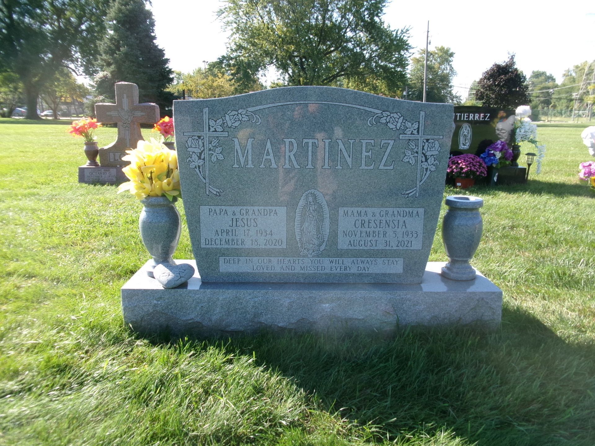 A gray granite headstone for the Martinez family in a grassy cemetery, flanked by vases with yellow and red silk flowers.