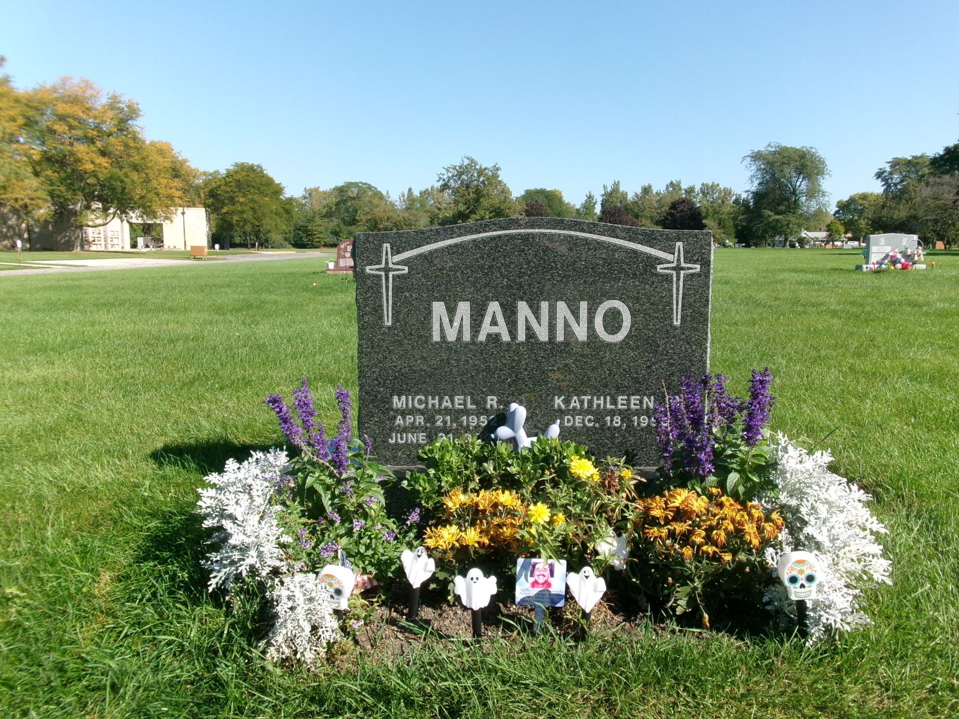 A dark granite headstone for the Manno family, adorned with purple and yellow flowers, sits in a sunny green cemetery.