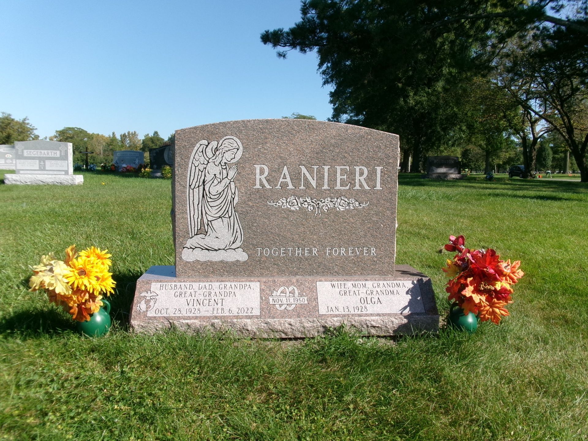 A granite Ranieri family headstone in a cemetery, featuring an angel engraving and floral arrangements on both sides.