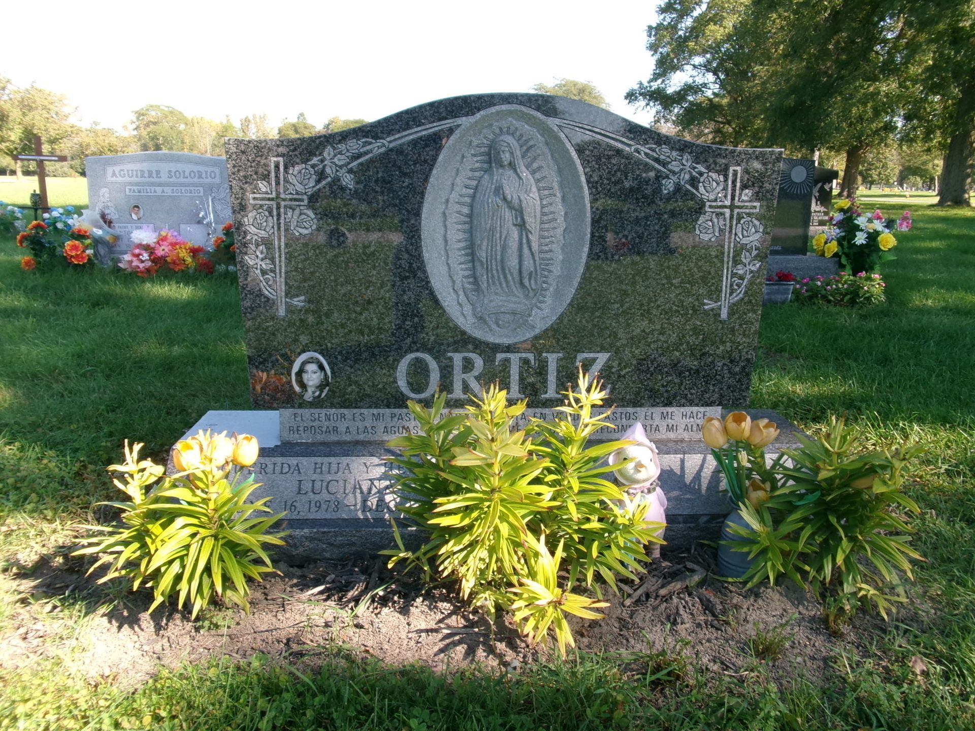 A granite cemetery monument for the Ortiz family, featuring an etched image of the Virgin Mary, set in a green lawn.