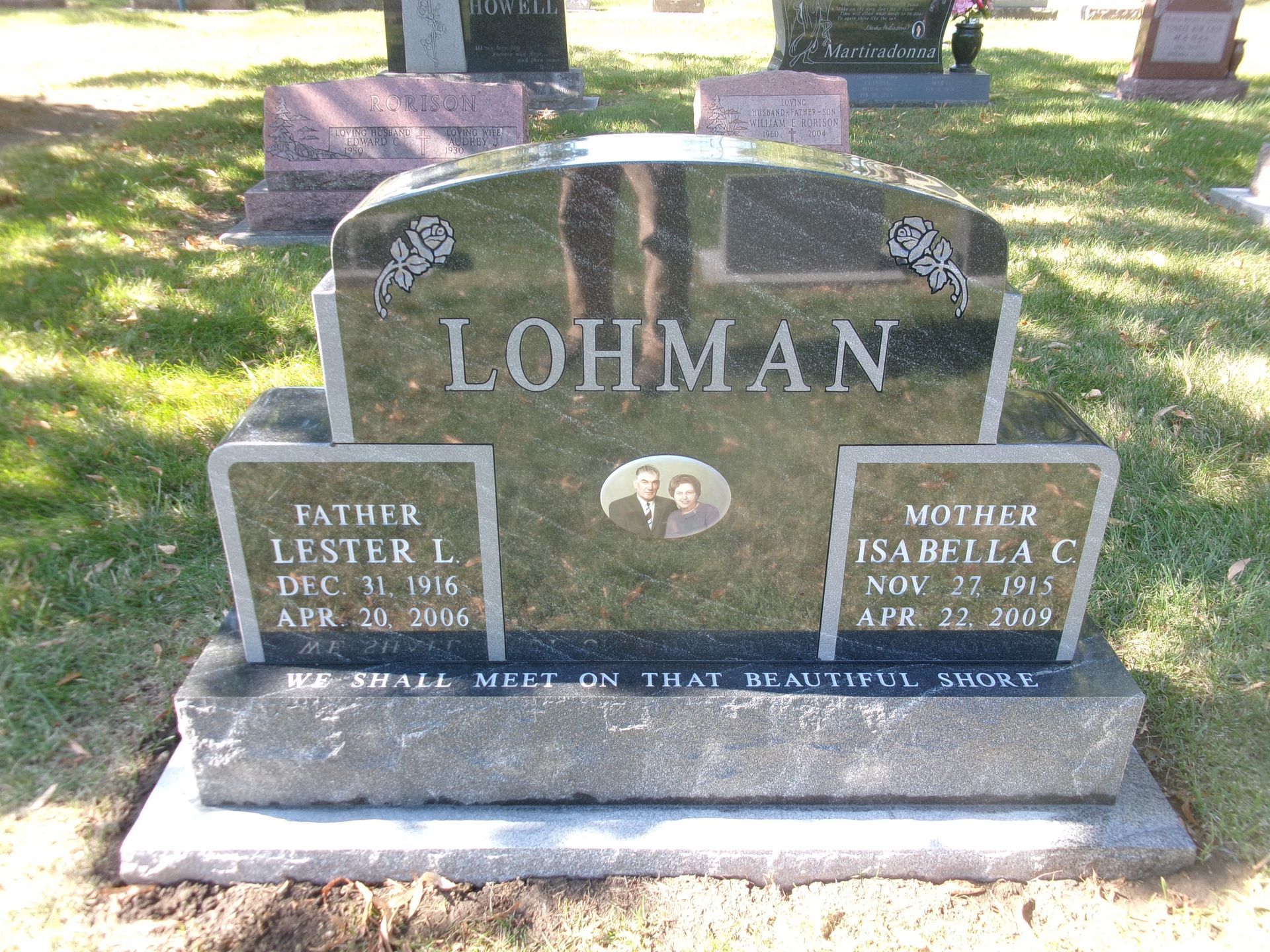 Black granite double headstone for Lester and Isabella Lohman, with a central photo, set in a sunny cemetery.