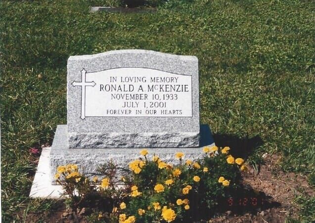 Gravestone of Ronald A. McKenzie with cross and dates 1933-2001, surrounded by yellow flowers and green grass.