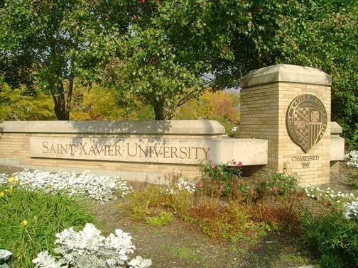 Sign for Saint Xavier University, with beige brick and stone and green foliage.