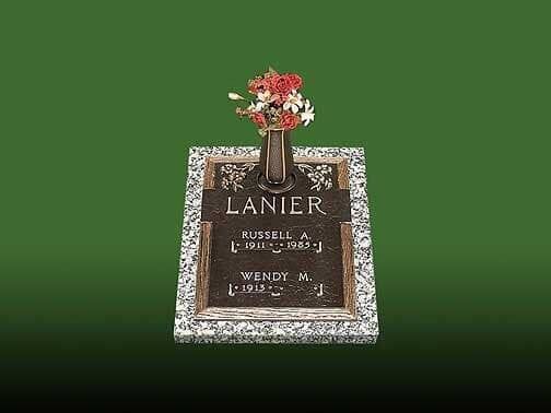 Bronze grave marker with flowers and cross, set in a stone base, on green grass.