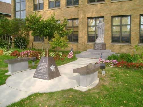Memorial with statue, benches, and flags in front of a brick building.