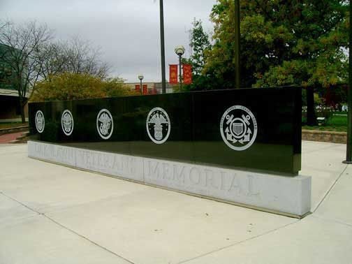 Black granite veterans memorial with service emblems, on a concrete base, outdoors.