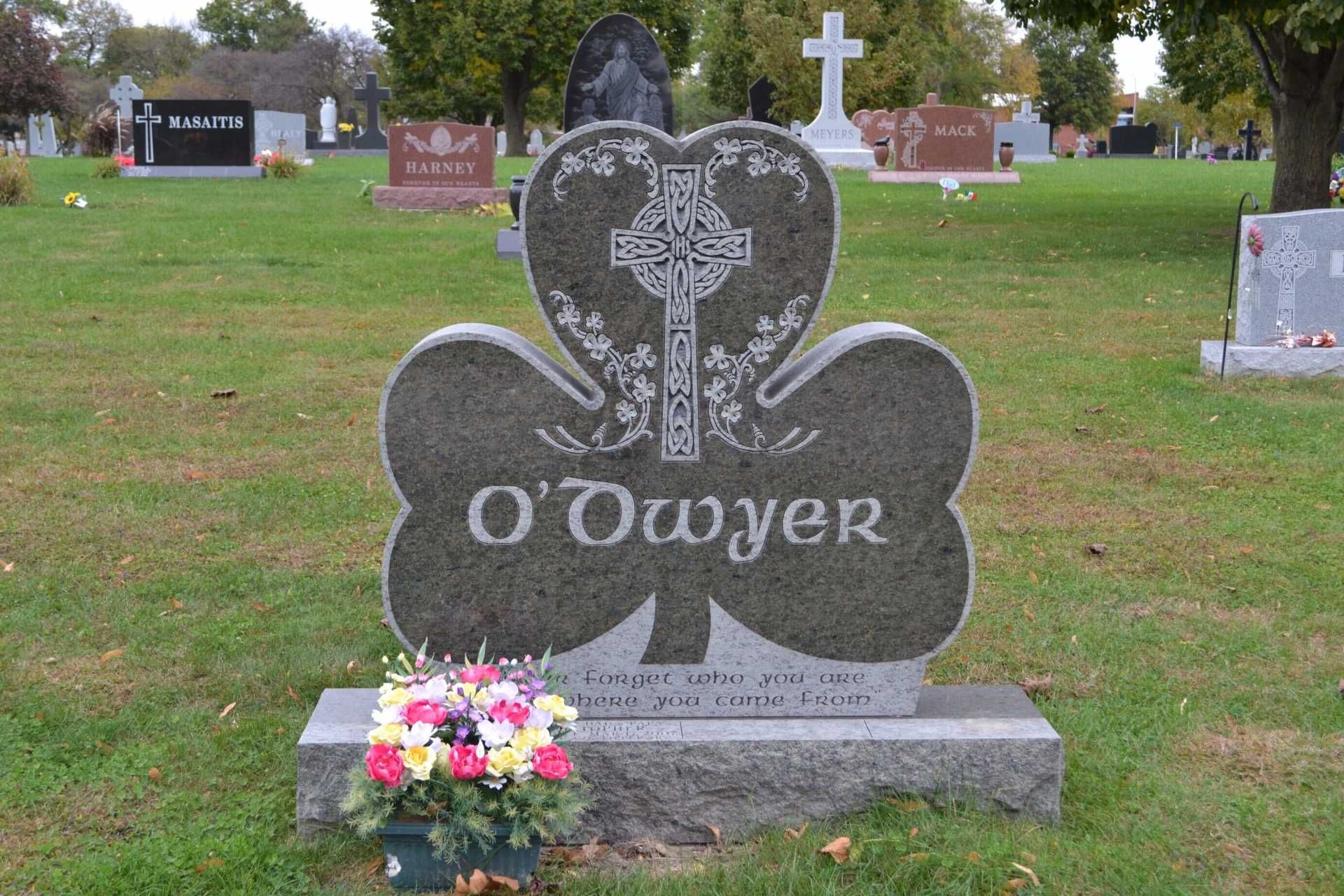 Clover-shaped tombstone with O'Dwyer inscribed, a Celtic cross, and a flower arrangement at a cemetery.