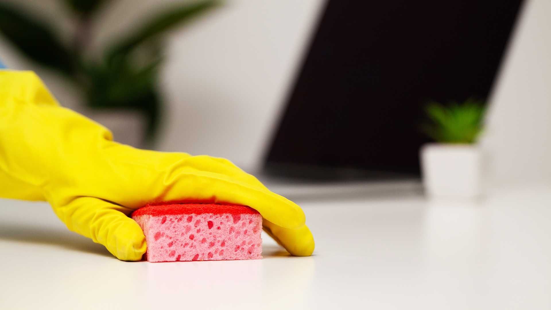 A person wearing yellow gloves is cleaning a desk with a sponge.