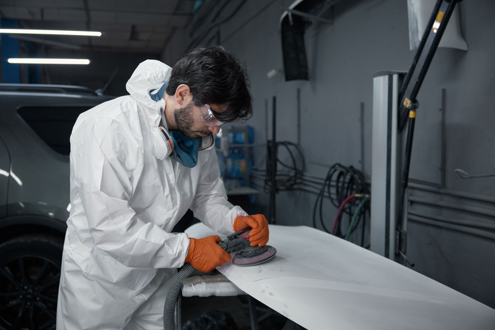 An auto body technician in a protective suit meticulously sands a car hood at a repair shop.