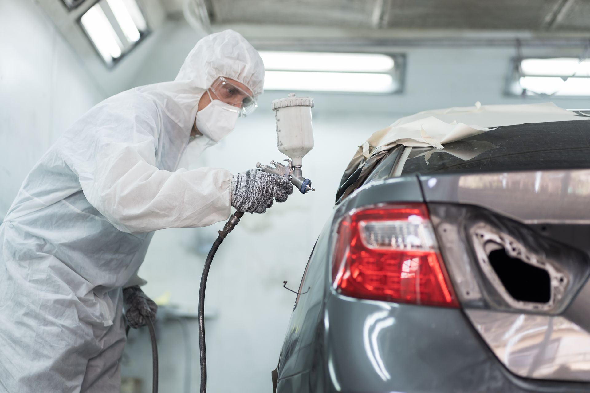 A mechanic is performing automotive spray painting with a spray gun on a vehicle at an auto shop.