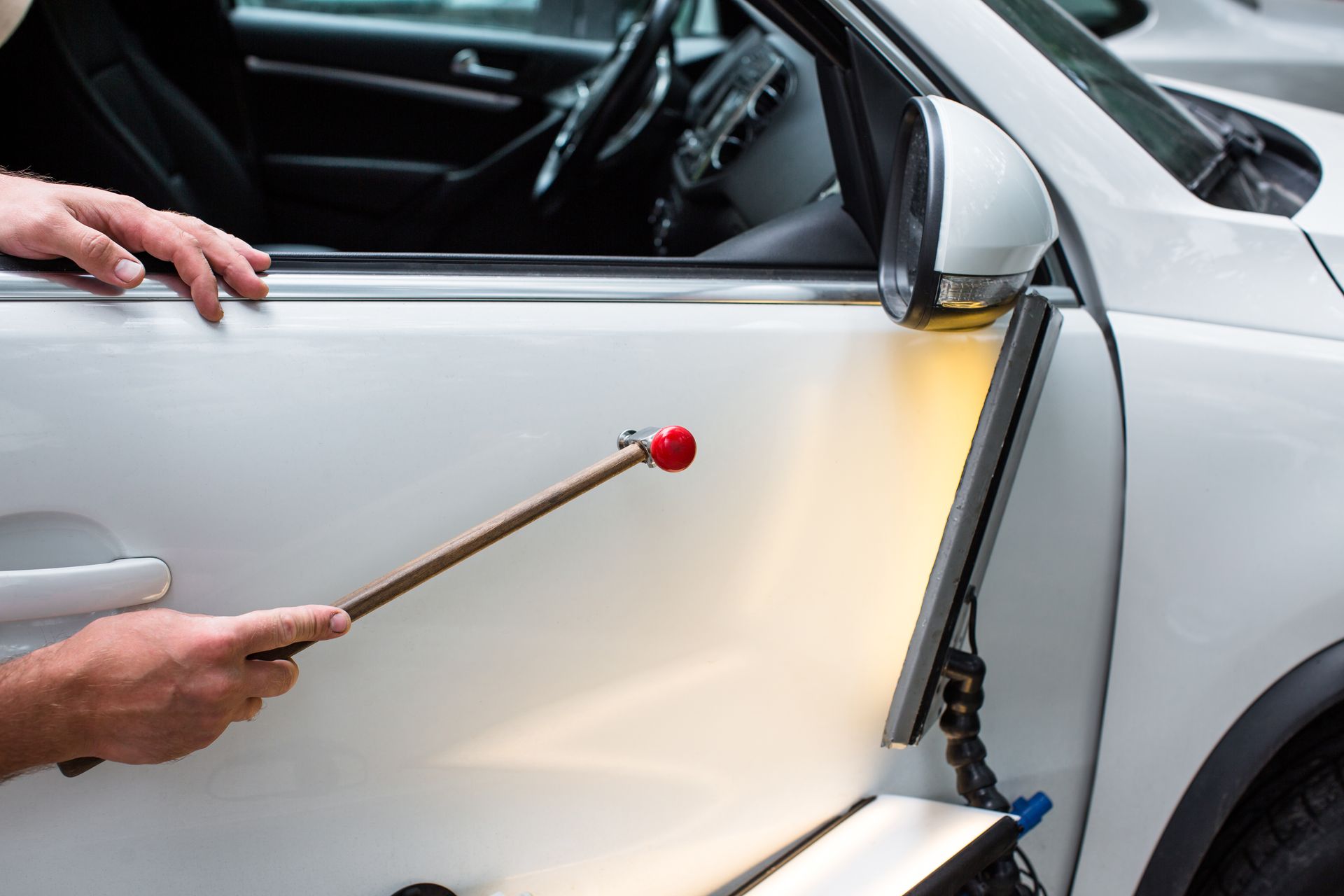 Technician uses a red-tipped tool for dent repair on a white car door under bright light.