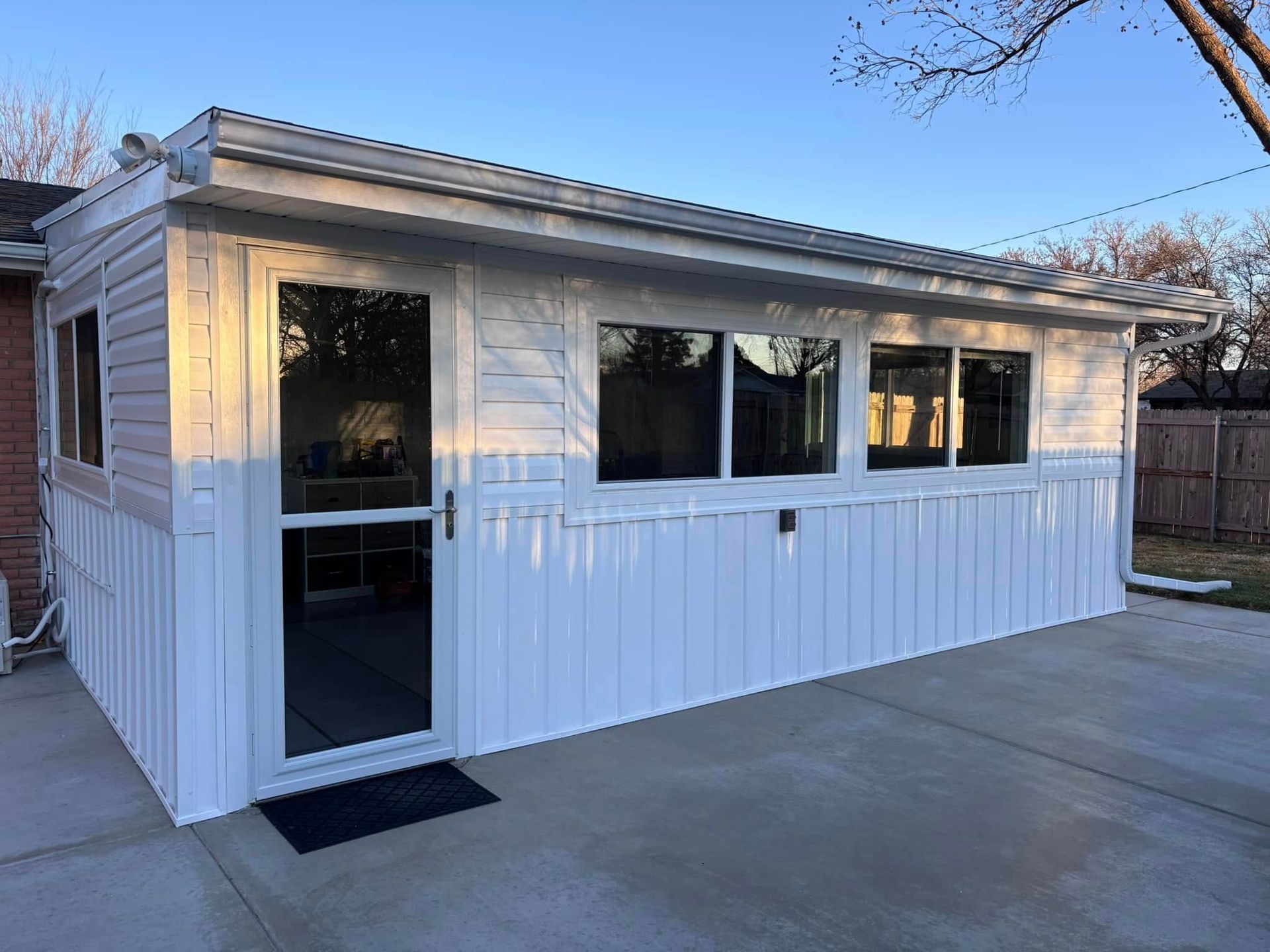 A white shed with a door and windows is in the backyard of a house.