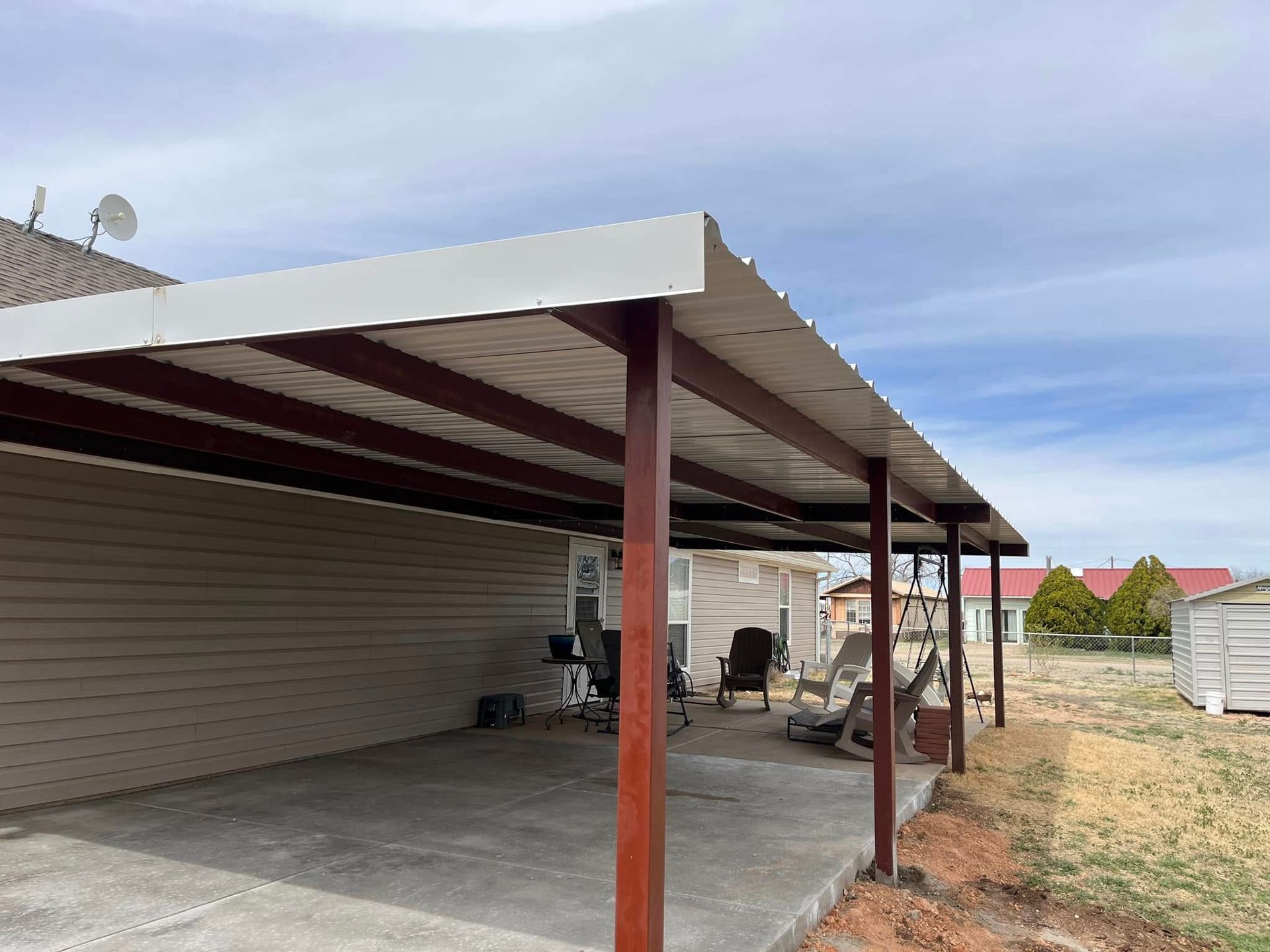A carport with a white roof and wooden beams is in the backyard of a house.