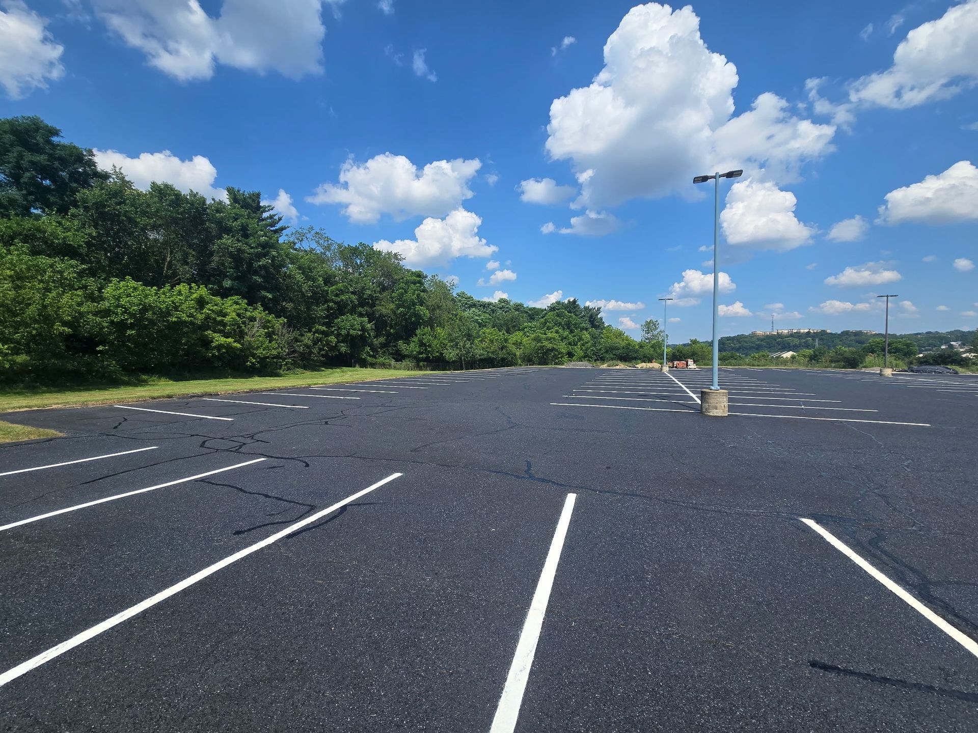 Empty parking lot under a blue sky with clouds, lined with white spaces and trees along the edge