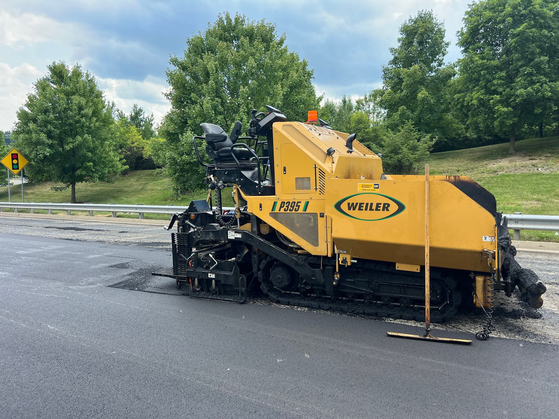 Yellow road paver laying fresh asphalt on a wet street, with trees and a cloudy sky in the background