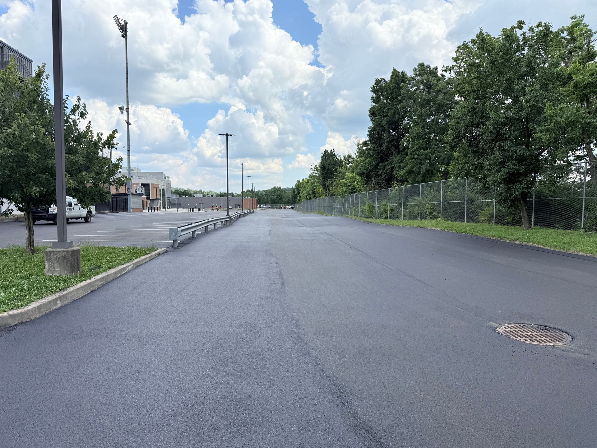 Empty asphalt road beside a chain-link fence and trees under a partly cloudy sky
