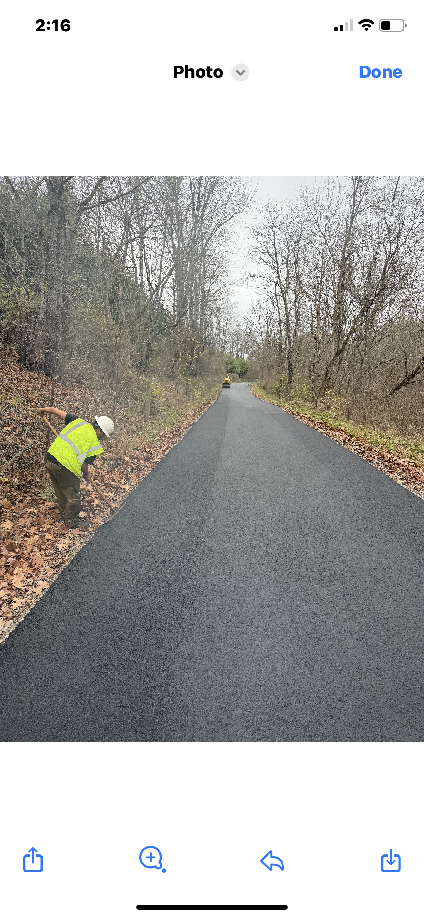 Person in a yellow jacket clearing leaves beside a paved road in a wooded area.