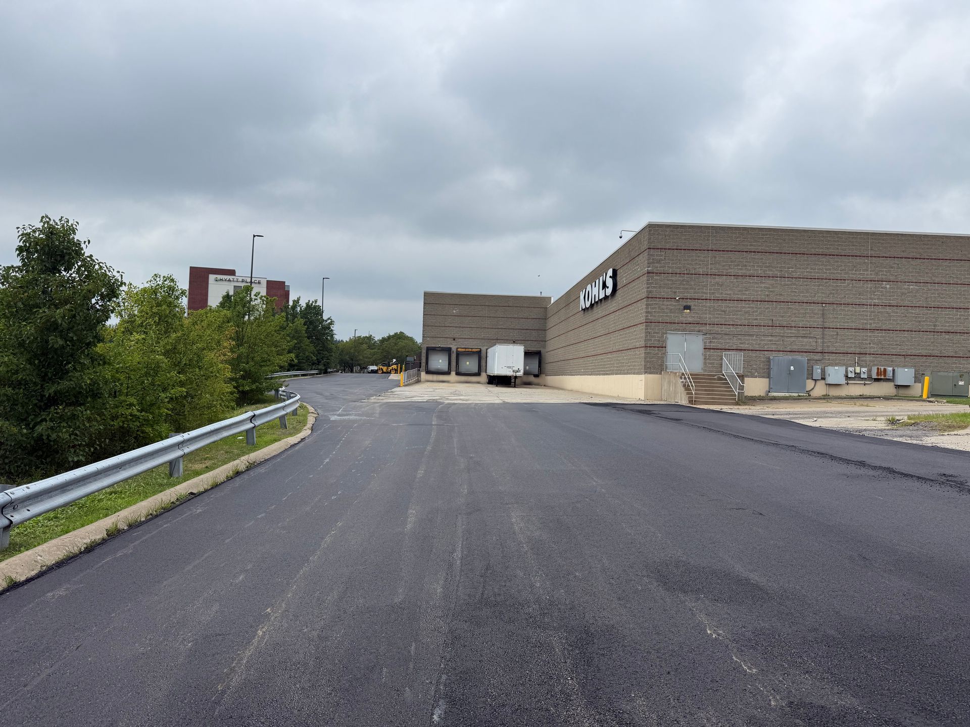 Industrial building and parking lot on a cloudy day, with a guardrail and trees along the road.