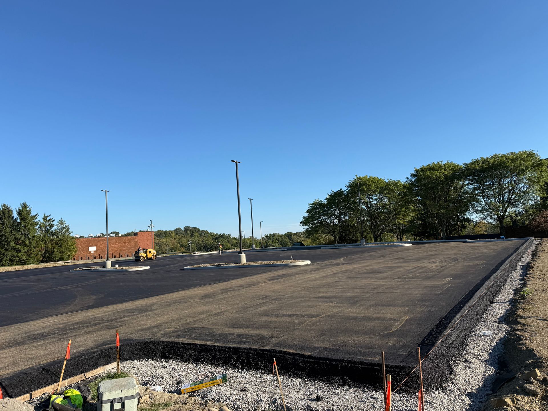 Freshly paved road surface on a construction site beside gravel and trees under a clear blue sky