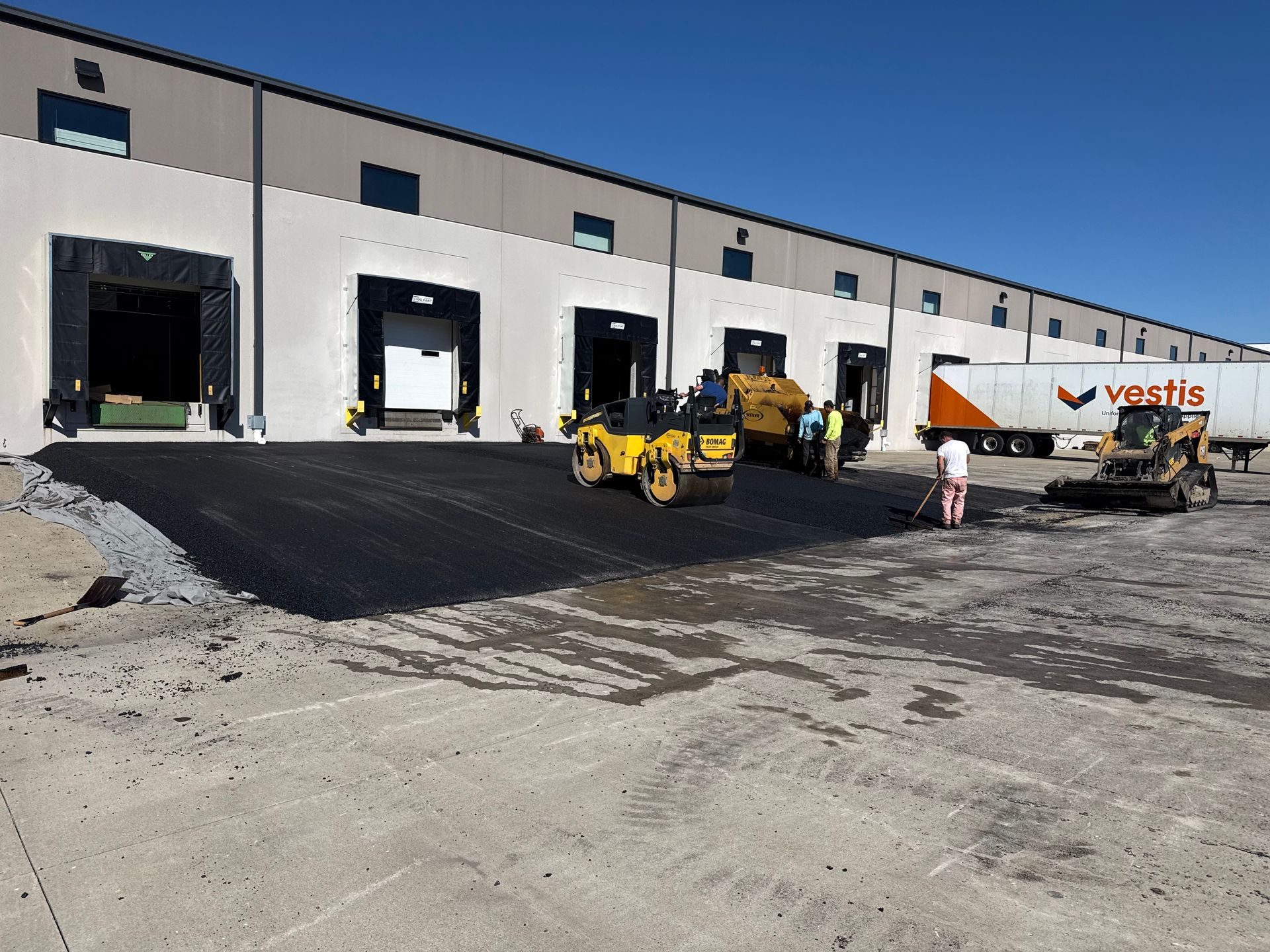 Industrial warehouse with open loading docks and construction equipment, including a yellow paver on black asphalt.