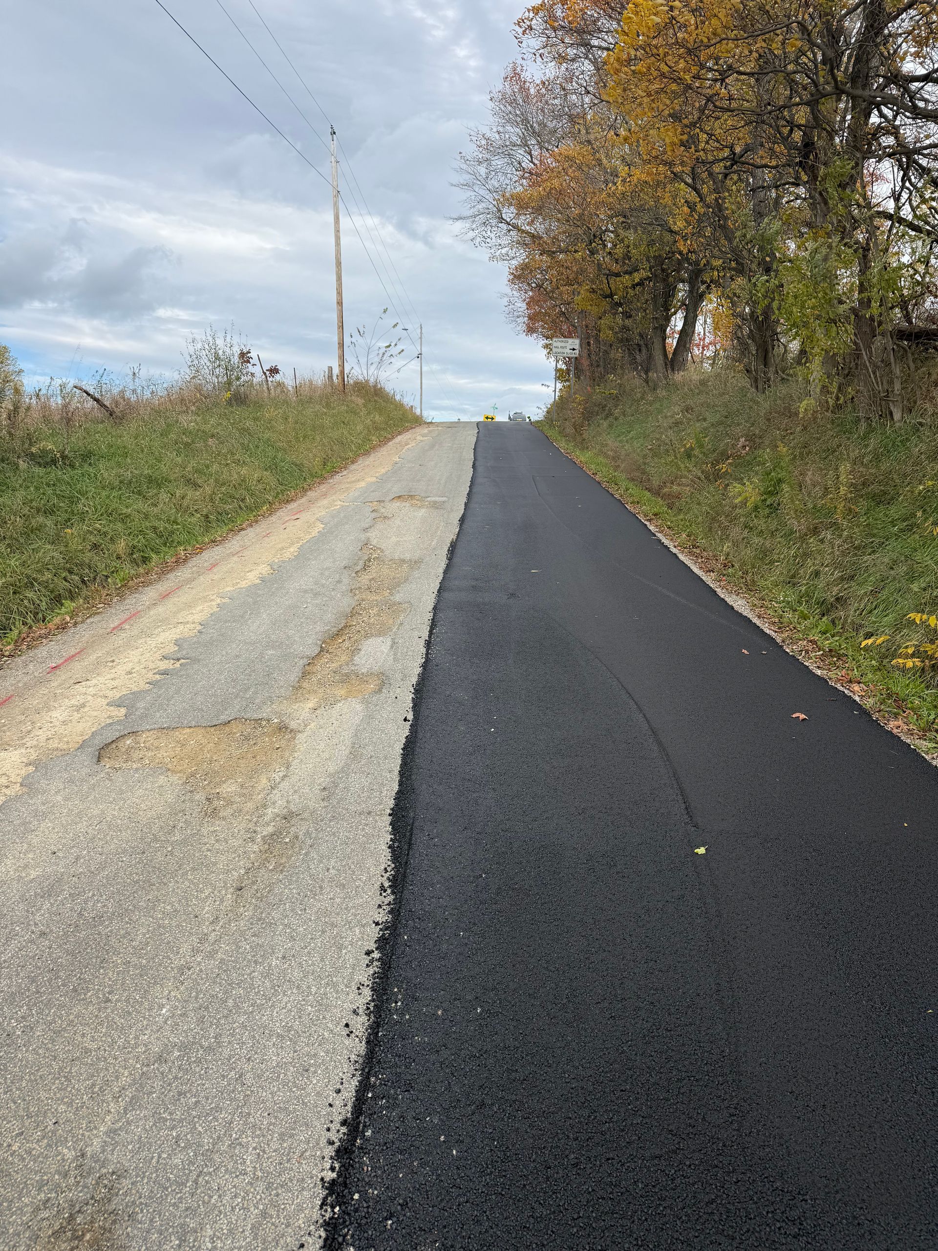 Newly paved rural road beside a gravel shoulder and autumn trees under a cloudy sky